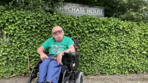 A man in a wheelchair wearing blue trousers, a green top and glasses. Behind him is a hedge and Chipchase House sign.