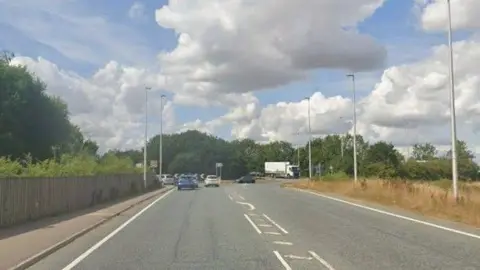 A view of the Wragby Road roundabout near Lincoln. The photograph is taken in the summer and the grass verge is going brown. A fence lines the other side of the road and tall lamp posts are either side of the carriageway.