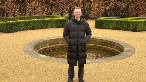 BBC/RICHARD EDWARDS A man, wearing a long black coat, standing alone in front of an ornamental pond. Evergreen, carefully trimmed hedges are behind him. He is smiling and has ginger hair.