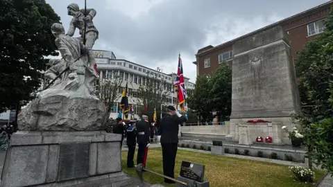Royal British Legion personnel salute the memorial which has several wreaths laid at the bottom. The memorial is a large oblong stone statue standing upright.A large stone statue of two army personnel is in the foreground. 