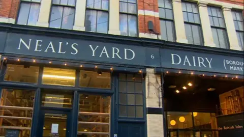 The outside of the store in Borough Market, has Neal' Yard Dairy written in white and blue sign with empty shelves in the front window and yellow bulbs hanging down on the right hand side of the store.