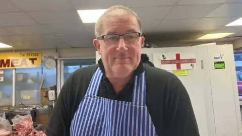 Lara King/BBC Market trader Gary on his meat stall in Freeman street market, he is wearing a blue and white vertically striped apron. He is standing in front of a fridge and packs of raw meat are on a counter behind him. 