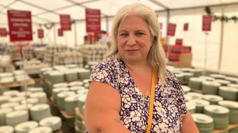 A woman with shoulder length hair and a blue and white floral top is looking at the camera. There are rows of crockery behind her