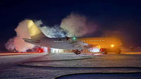 A large plane sits on the snow in the dark with clouds of vapour around it