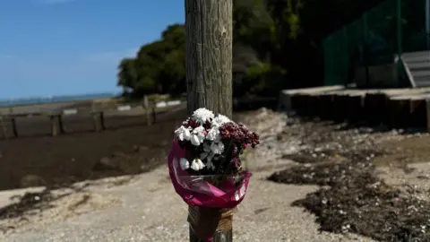 BBC A bunch of flowers tied to a wooden stump on the beach at Binstead. The sea is visible on the left and the sky is blue.