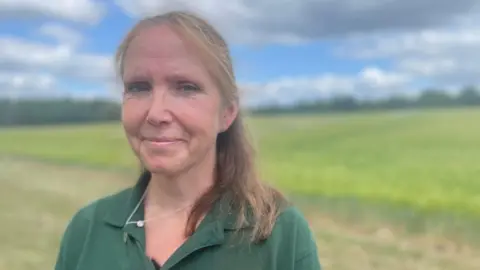 Malcolm Prior/BBC Jo, with tied-back strawberry-blonde hair and a green polo shirt, stands in front of a farmer's barley field in the sunshine, with a stretch of cloud across an otherwise blue sky.