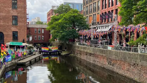 BBC / Rob Oxley A canal runs between red-brick buildings decorated with rainbow flags, with crowds gathered along a waterside terrace.Reflections ripple on the water below as people socialise outside bars and venues lining the canal.
