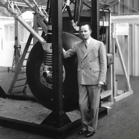 George Dowty A black and white photo from 1942 of Sir George Dowty, in a very dapper suit and tie, with his hand resting on the testing rig of the Dowty undercarriage, fitted to the Lancaster aircraft
