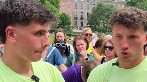 The brothers are on the left and right of the photo and both wearing a green T-shirt. A group of people is behind them and a building is in the background.