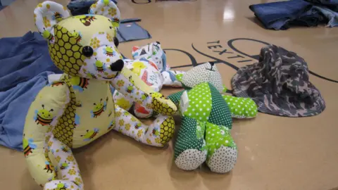 Alex Pope/BBC Three cloth made teddy bears on a table, with a hat next to them. Denim items are also on the table. The table is large and cream in colour, with writing in the middle. 
