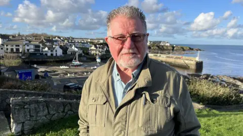 A man with short grey hair and glasses, wearing a blue polo shirt and beige jacket, He is stood on a grassy verge, with a view over a harbour behind him,