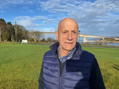 A man with a blue body warmer and navy jumer, infront of a green grassy area. In the background, blue skies are disrupted by clouds, with a bridge and trees also present. 