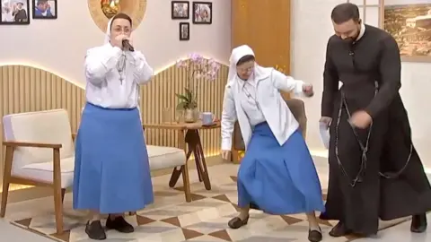 A nun beatboxing into a microphone with another nun and a priest dancing beside her