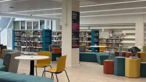 Reading Borough Council Image of a library with rows of books and yellow, blue and grey chairs and sofas. 