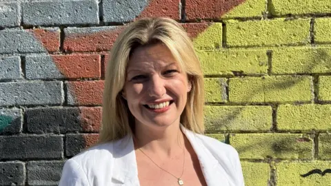 Blonde haired woman wearing a white blazer and a necklace.  She is smiling and standing by a brick wall that is grey but painted in part in yellow and red.