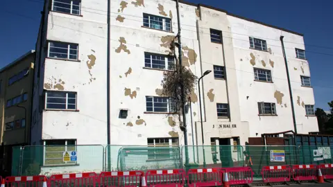 A dilapidated four-storey building. White paint is flaking off it, with windows lining he side. There are red and white striped barriers in front of it and metal railings. 