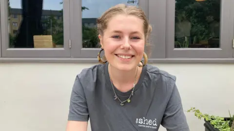 Tilly Yong is sitting outside the Shed cafe in Taunton.  She is wearing a grey T-shift with The Shed written on it.  She is also wearing a necklace.
