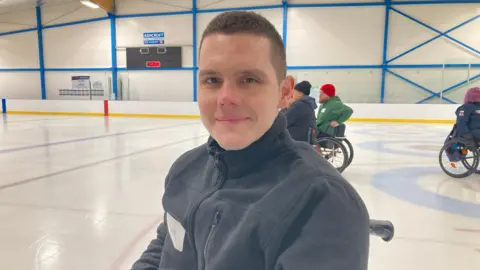 Tom Williams/BBC Jake McHale is looking at the camera and smiling. He is on the ice in his chair, wearing a grey jacket. People in wheelchairs can be seen on the rink behind him