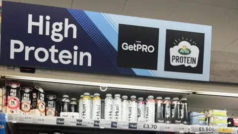 Uma prateleira de supermercado com bebidas de Christine Roe e produtos de alta proteína com iogurte.