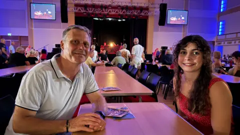 A side-on picture of an older man and a younger woman smiling at a busy music event. The pair sit at a bench-style table. Surrounding them are people sitting at neighbouring tables and others dancing. A stage with bright lights and screens can be seen in the background. The man wears a light-blue polo shirt with black detailing on the arms and collar, the woman a red polka-dot dress and has brown curly hair.