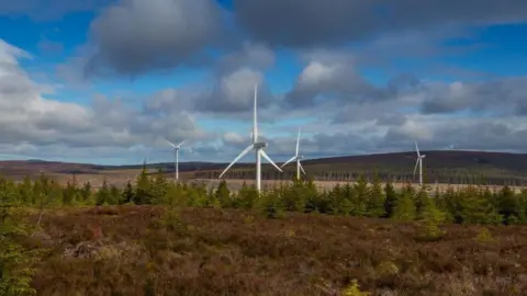 Wind turbines among trees, with hills and a cloudy blue sky