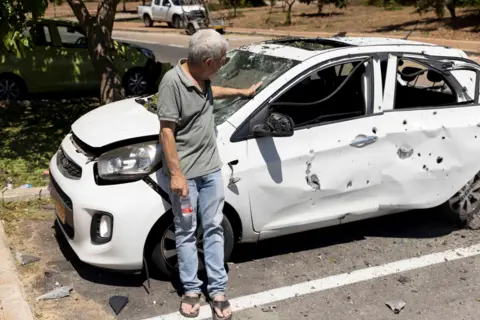 Getty Images A man stands next to his wrecked car which has been hit by shrapnel 