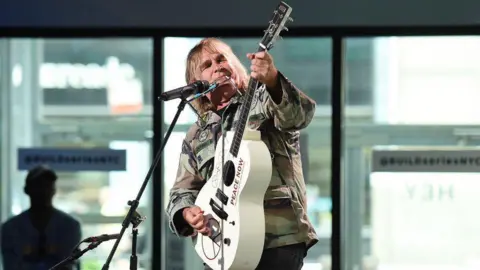 Mike Peters who has long blonde hair and is wearing a khaki camouflage jacket. He is stood at a microphone stand and is holding a cream guitar which he is playing and looking at. Behind him are some large windows and a man wearing a baseball cap.
