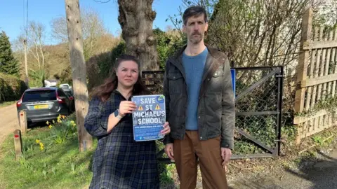 Claudia Parker and Geoff Black look at the camera. They are holding a sign which says 'Save St Michael's School.'