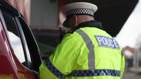 A police officer wearing a black-and-white police hat and yellow high-vis jacket stands next to a dark red vehicle's driver-side window. 