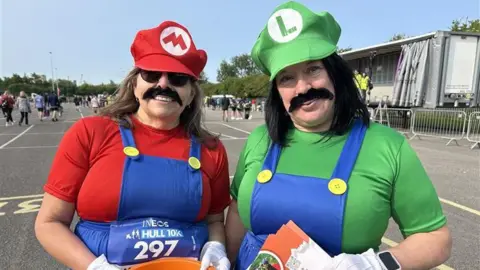 Gill wears a red t-shirt and cap and Nicola wears a green t-shirt and cap. The caps have M and L on them and they both wear thick black moustaches.