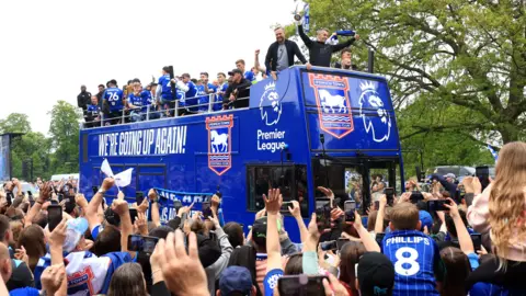 PA Media A blue open top double decker bus travels through a crowd. Dozens of people wearing blue football shirts look on and cheer. There are men at the top of the bus looking down and similarly cheering.