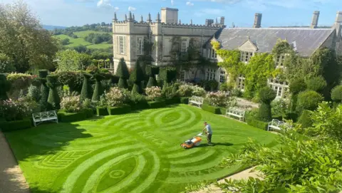 A stately home with a neatly cut lawn and ornate benches dotted around.