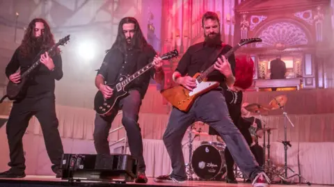 Ewan Cawood Two long haired men with guitars and one short haired man with a beard play guitars in front of a red haired wearing a black blouse with a white collar. In the background a man plays a church organ.