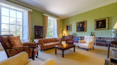 The Armstrong Family The picture shows a large, elegant drawing room. The space is bright and airy, with two tall sash windows on the left-hand wall letting in natural light and giving views of trees outside. The walls are covered in green patterned wallpaper, topped with detailed white cornicing that runs around the ceiling.
The room is furnished in a traditional style. In the centre is a dark wooden coffee table, surrounded by several upholstered armchairs and sofas in warm, muted tones, including creams and deep reds with patterned fabric. 