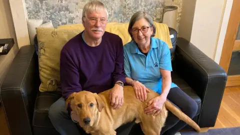 Luke Deal/BBC Colin and Anne Daultrey sit on a brown leather sofa and smile at the camera. They both have grey hair and sat around their legs is Karter the puppy. He is a golden colour. The couple smile at the camera.