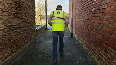 A man in a high vis jacket walks away from the camera down an alleyway between two brick walls