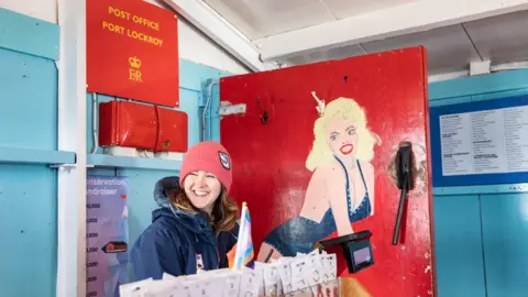 UKAHT Natalie Corbett smiling inside the post office. She is wearing a navy coat and a red beanie hat. There is a red Post Office sign behind her and key rings on a stand in front of her.