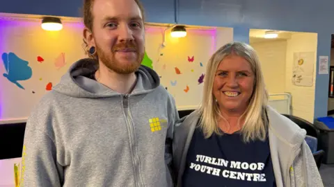 A man with ginger hair smiling whilst wearing a grey hoody with yellow writing on stood next to a woman with blonde hair smiling and wearing a t-shirt that reads Turlin Moor youth centre.