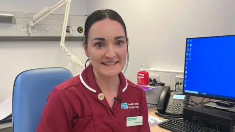 Stephanie Todd is pictured in a clinical environment. She is smiling and looking toward the camera. She has long brown hair tied up and is dressed in maroon medical scrubs. She is pictured in a clinical environment, behind her is a busy desk and a computer screen.