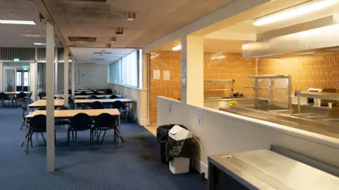 PA Media An empty dining room with tables and chairs laid out on a blue carpet. There is a serving area to the right.
