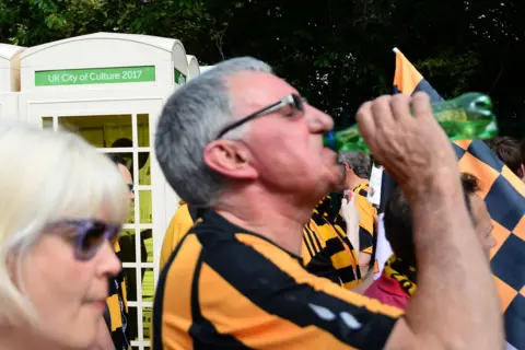 Getty Images A woman and a man dressed in a black and amber football shirt pass close to the camera with cream phone boxes behind them. The man is drinking from a green water bottle. Other fans dressed in the same shirts stand behind him. A black and amber flag is waved. The phone boxes include a green sign reading: UK City of Culture 2017.