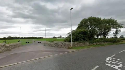 Google Google maps street view of a gate to a field. There's a road and a brick wall. There's a a few trees by the hedge.