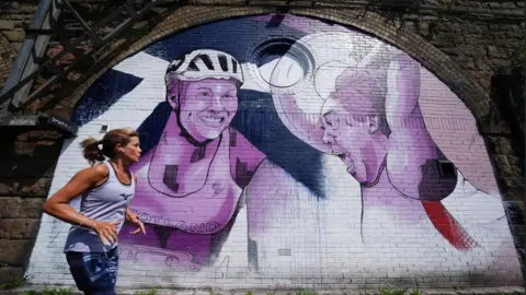 PA Media A runner runs past a white, purple and blue mural featuring Team Scotland stars.
