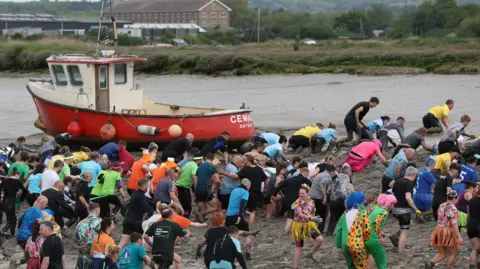 Chris Radburn/Fixedpoint Media A large crowd of people dressed in colourful outfits crawl through thick mud on a river bank. To the right Mr Rugg is also on his hands and knees, dressed as a bright pink flamingo.