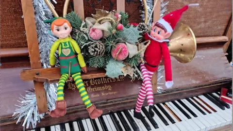 North East Lincolnshire Council Green elf on the left of a Christmas themed decoration and red elf on the right. Both sitting on the music stand of an old piano