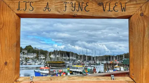 Aileen Gilies Harbour scene framed by a wooden viewing panel, looking out over fishing boats and masts beneath a cloudy sky.