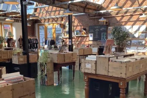 Just Dropped In Just Dropped In record store, in Coventry's Fargo Village. It has crates of vinyl records on tables, with the shop also selling clothing which is on rails. The shop is decorated with plants. 