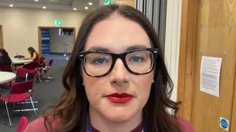 A woman with long brown hair and glasses looks at the camera. She is wearing a maroon top and is standing next to a brown door.