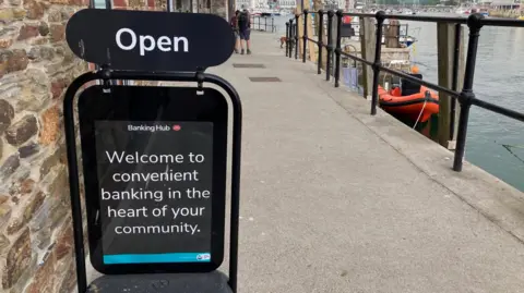 BBC A sign that reads "welcome to convenient banking in the heart of your community" on a quayside pavement outside a banking hub.