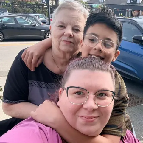 Natalia Sidorska Natalia is hugged by her son and mother. They are outside in a car park. 